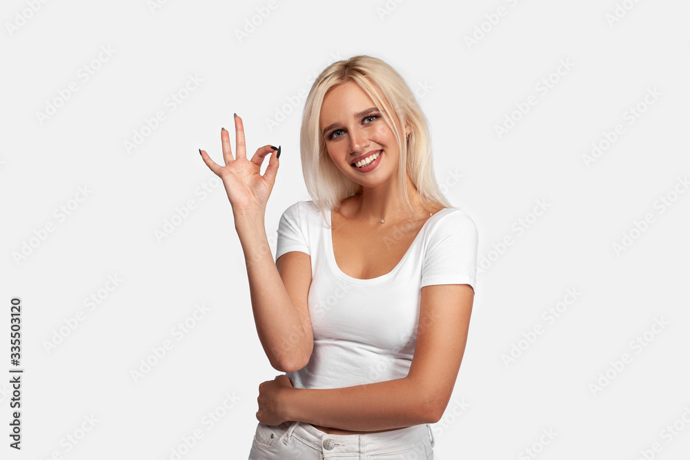 Young beautiful blonde woman wearing casual t-shirt standing over isolated white background holds an okay gesture with hand. Ok sign. Mockup.