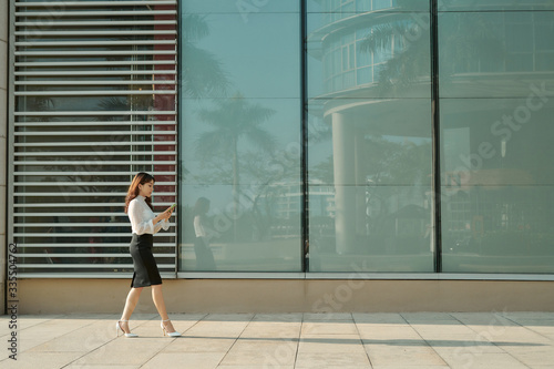 Full length side portrait of a young woman walking and sending text message on cell phone