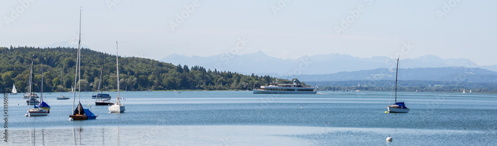 Herrsching, Bavaria / Germany - Aug 11, 2019: Panorama view of Lake Ammersee with sail boats and a steam boat. Far in the distance / background the bavarian alps. Amazing spot for nature lovers.