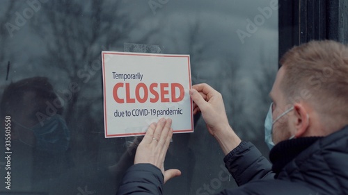 Caucasian male wearing medical mask puts a Temporary closed due COVID-19 pandemic sign on a window. Coronavirus pandemic, small business shutdown photo