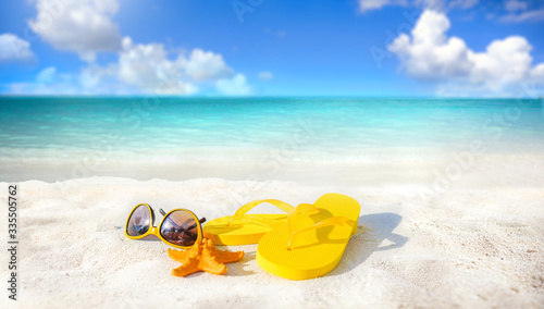 Fototapeta Naklejka Na Ścianę i Meble -  Concept of summer beach holiday. Beach accessories - sunglasses, starfish, yellow flip-flops on sandy tropical beach against blue sky with clouds on bright sunny day. Beautiful colorful image.