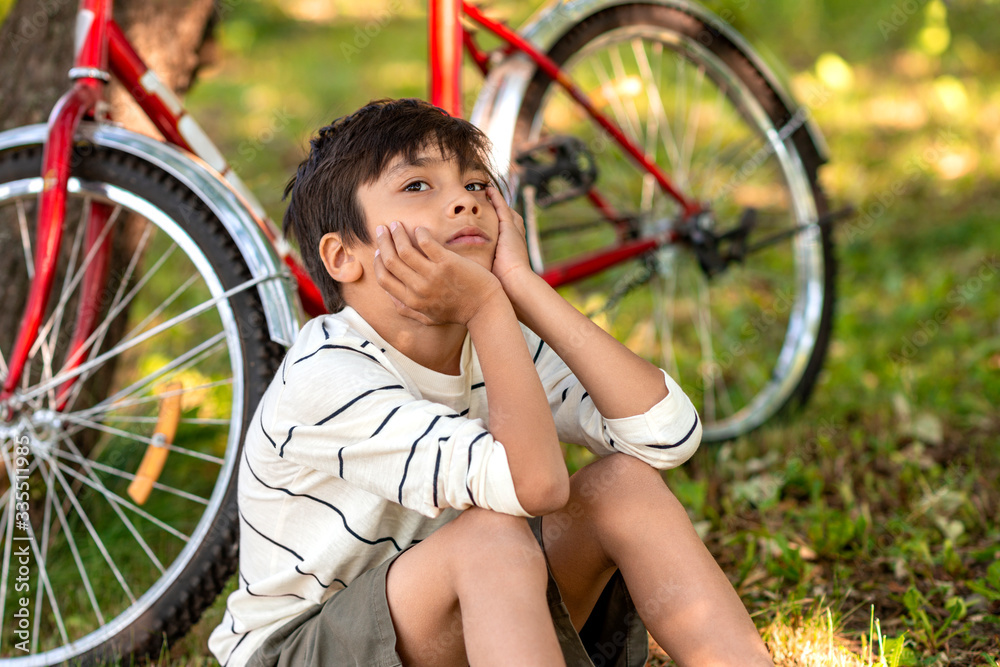 Obraz premium A young boy sits on the grass in the garden and dreams of something. A red bike is standing by a tree, a book is lying on the grass