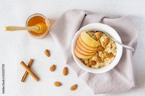 Oatmeal porridge with honey  apple  and almond nuts in a bowl served on grey napkin. There is jar of honey  cinnamon  and almond nuts. Vegan or vegetarian breakfast. Grey background  top view. 