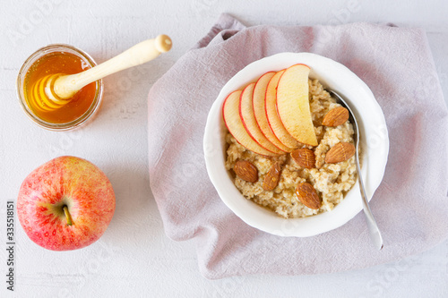 Vegan or vegetarian breakfast. Oatmeal porridge with honey, apple, and almond nuts in a bowl served on grey napkin. There is jar of honey and apple near it. Grey background. 