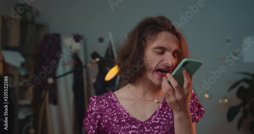 Close up view of angry transgender man in shouting fuck you into smartphone loudspeacker. Drunky transgender yelling on friend while standing at home in fashionable dress with sequins. photo