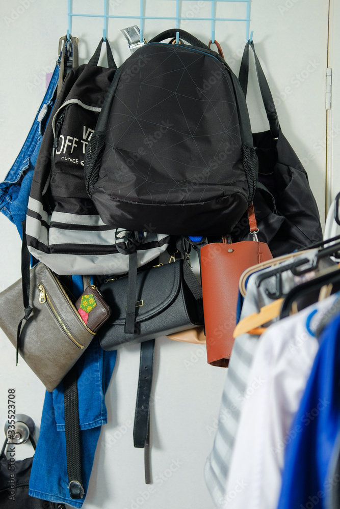Collection of multiple backpacks, handbags and a pair of jeans stacked and hanging on a rack behind the door at home. Collection of multiple backpacks, handbags and a pair of jeans stacked and hanging on a rack behind the door at home.