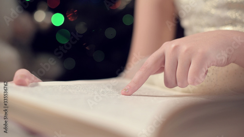 Wallpaper Mural Close-up soft focus the hand of a little girl leads along the lines of a book while reading a New Year's fairy tale amid the lights of a Christmas tree. The concept of a fabulous mood Torontodigital.ca