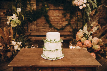 White wedding cake decorated with green leaves and stands on a wooden table in the loft