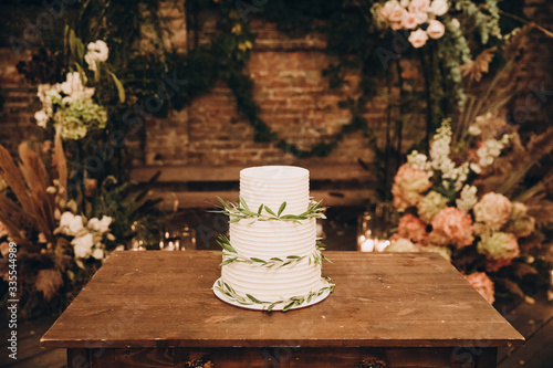 White wedding cake decorated with green leaves and stands on a wooden table in the loft