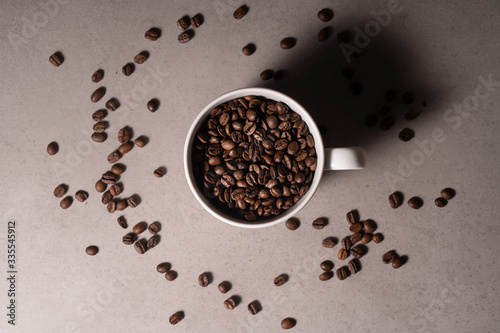 Zdjęcie białego kubka z ziarnami kawy na szarym tle, dookoła rozsypane ziarna kawy