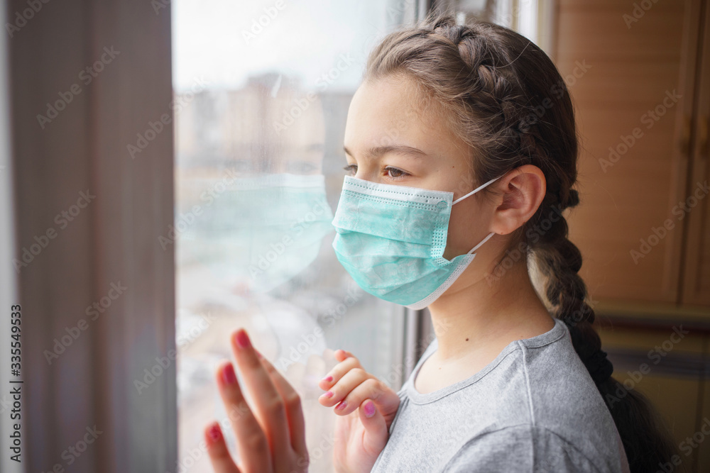 a girl in a medical mask looking through window. at the quarantine caused by coronavirus