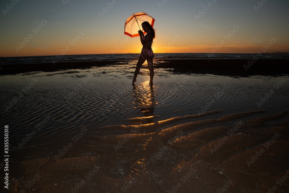 Obraz premium Silhoutte of a pretty senior Asian female in a swimming suit standing and hold umbrella posing on the beach..