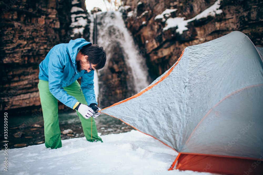 Young professional male tourist is preparing to camp in the mountains near the river in winter. White waterfall and beautiful texture of rocks on background. Travel and trekking concept.