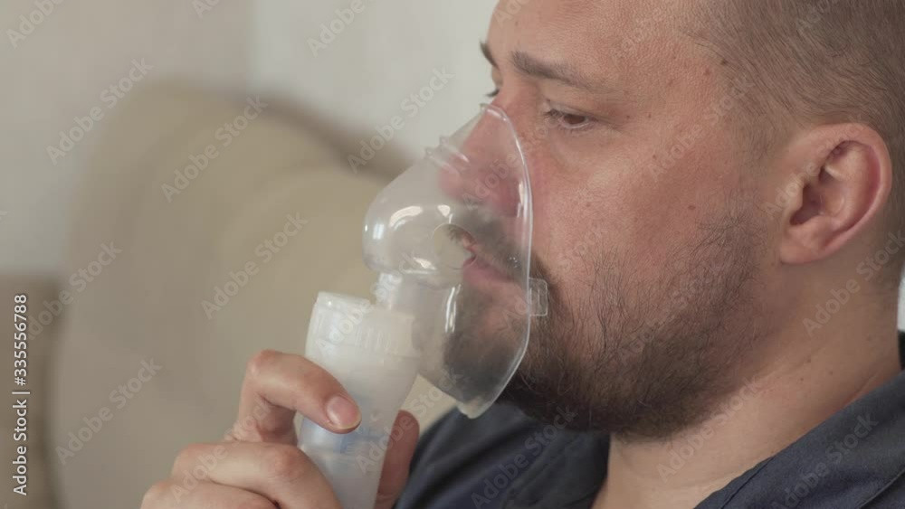 Middle-aged European man with a beard makes inhalation sitting on the couch and coughs Middle-aged European man with a beard makes inhalation sitting on the couch and coughs