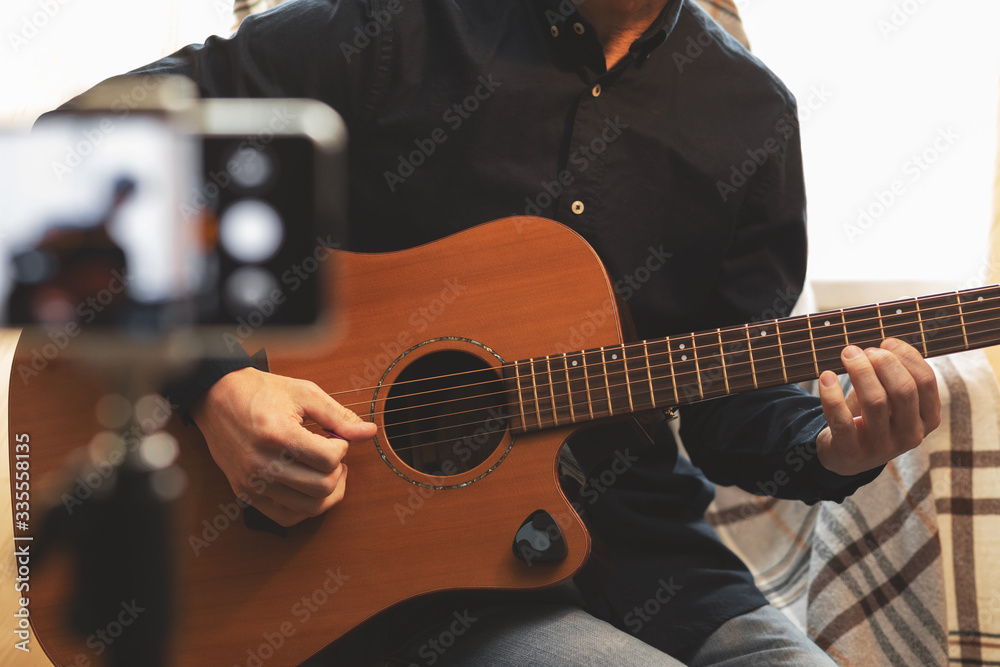 Obraz premium Man with acoustic guitar in a shirt and cap plays live staying home for the audience in front of the smartphone camera during quarantine and forced self-isolation due to coronavirus