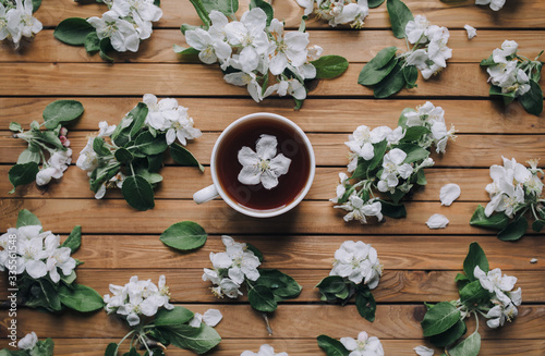 White cup of tea with petals of an apple tree on a wooden background, view from above.
