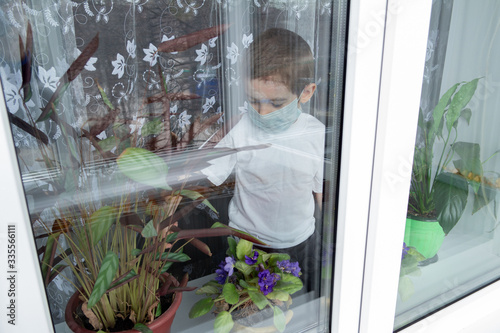 A boy in a mask sits at home on a windowsill and takes care of indoor plants and flowers. Concept of classes in a mode of self-isolation during a pandemic coronavirus or home treatment of the disease.