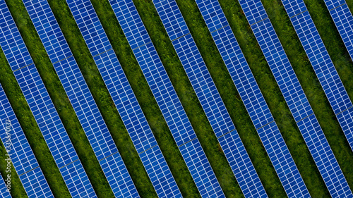 Aerial drone photo of a photovoltaic solar cell farm. Dark blue solar panels against a contrasting green grass field.
