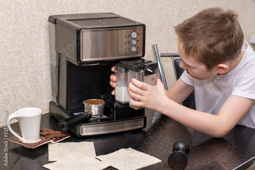 7 year old boy in a white T-shirt is preparing a hot cappuccino in a home coffee machine. sprinkled with cinnamon