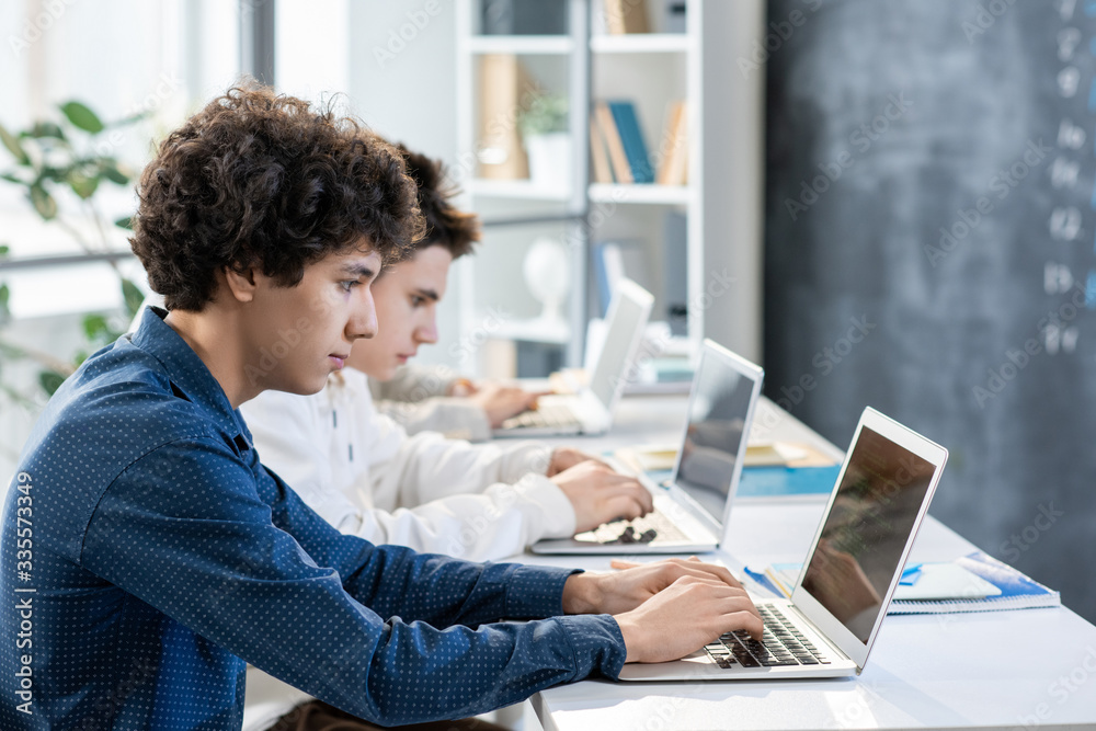 Side view of two guys sitting by desk in row in front of laptops Side view of two guys sitting by desk in row in front of laptops
