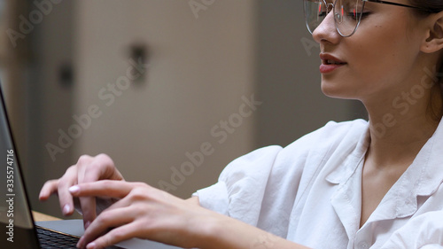 Fototapeta Naklejka Na Ścianę i Meble -  Freelancing and remote work. Young woman with glasses typing a letter on a laptop,