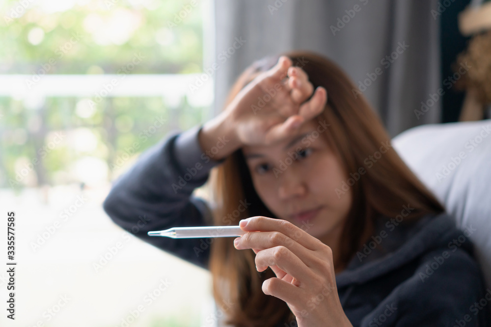 Young sick woman with fever checking her temperature with a thermometer at home. The concept of disease and ill Young sick woman with fever checking her temperature with a thermometer at home. The concept of disease and ill