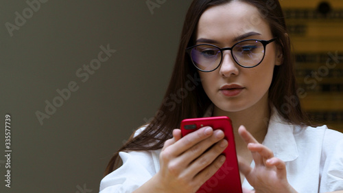 young girl with glasses uses a smartphone, close- up. The student holds in his hands the red phone