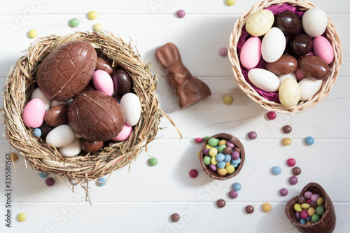Photography Top view of Chocolate eggs on nest, chocolate bunny, easter almonds and sweets on white wooden table