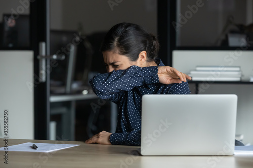 Sick young indian businesswoman working on computer in office, suffering from flu grippe corona virus symptoms, coughing in elbow. Unhealthy mixed race girl sneezing, feeling unwell at workplace.