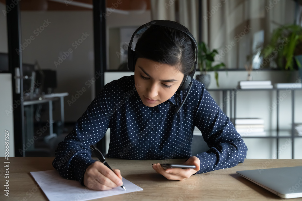 Focused young indian woman wearing earphones, holding smartphone in hands, writing notes on paper. Smart student passing exam or studying remotely, listening audio lecture in mobile application. Focused young indian woman wearing earphones, holding smartphone in hands, writing notes on paper. Smart student passing exam or studying remotely, listening audio lecture in mobile application.
