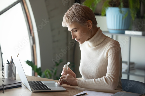 Focused responsible young 30s woman wiping computer keyboard and touchpad with antibacterial liquid, working alone distantly from home, preventing stop spreading coronavirus or dangerous infection. photo