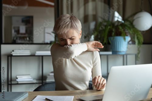 Photos Front view young female manager with short haircut coughing or sneezing in elbow while working on laptop in office room