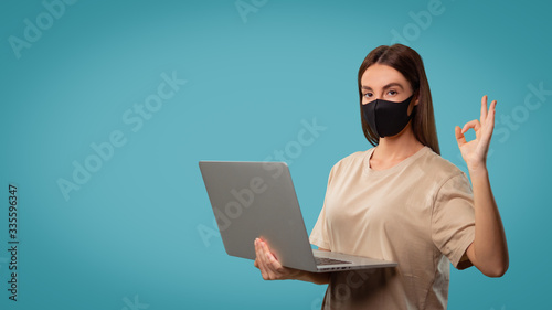 Woman in dust mask shows ok sign, she holds computer isolated over blue background. photo