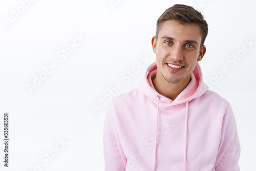 Close-up portrait of handsome blond man with blue eyes, smiling broadly camera with happy expression, showing positive feeling and upbeat attitude, standing in pink hoodie white background