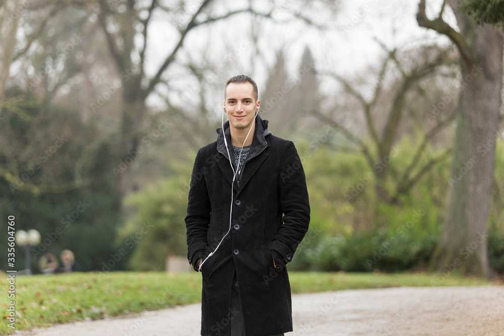 Young Man Smiling in Park Wearing Coat and Listening to Earphones Young Man Smiling in Park Wearing Coat and Listening to Earphones