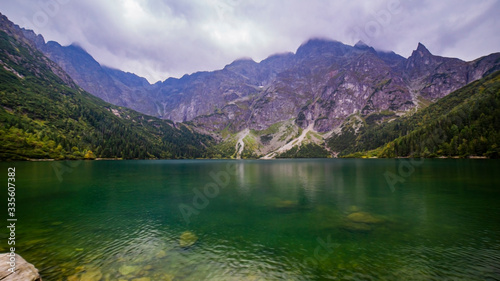 Scenic view of foggy mountains cover by dark clouds and green forest with a reflection in a lake. Stony shore. Morskie Oko. Marine Eye. High Tatras  Zakopane  Poland Concept of nature and tourism