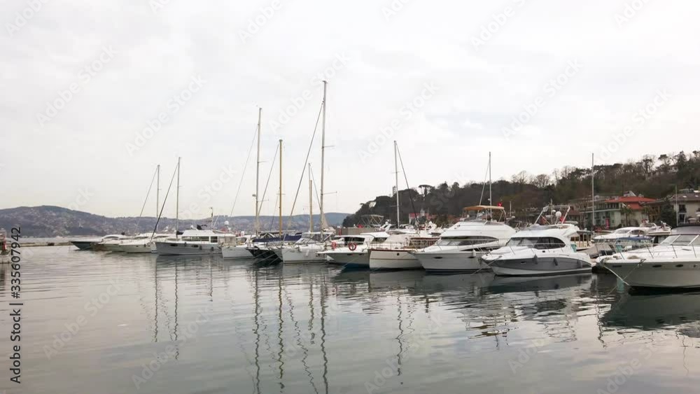Cloudy foggy day. Private yachts and sailing boats with lowered sails standing on pier in bay. Reflections in water. People walk along promenade. Turkey, Istanbul, Tarabya, March 2020. Cloudy foggy day. Private yachts and sailing boats with lowered sails standing on pier in bay. Reflections in water. People walk along promenade. Turkey, Istanbul, Tarabya, March 2020.