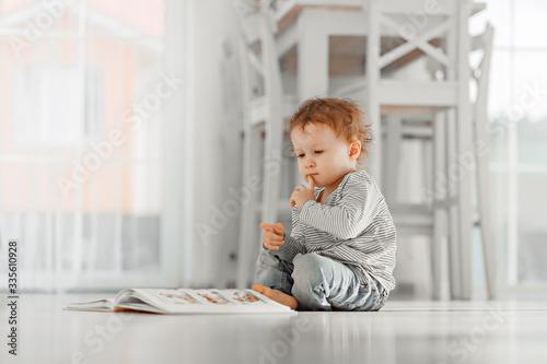 A little girl about 2 years old. In a light interior of neutral colors, reads a big book, backlight.