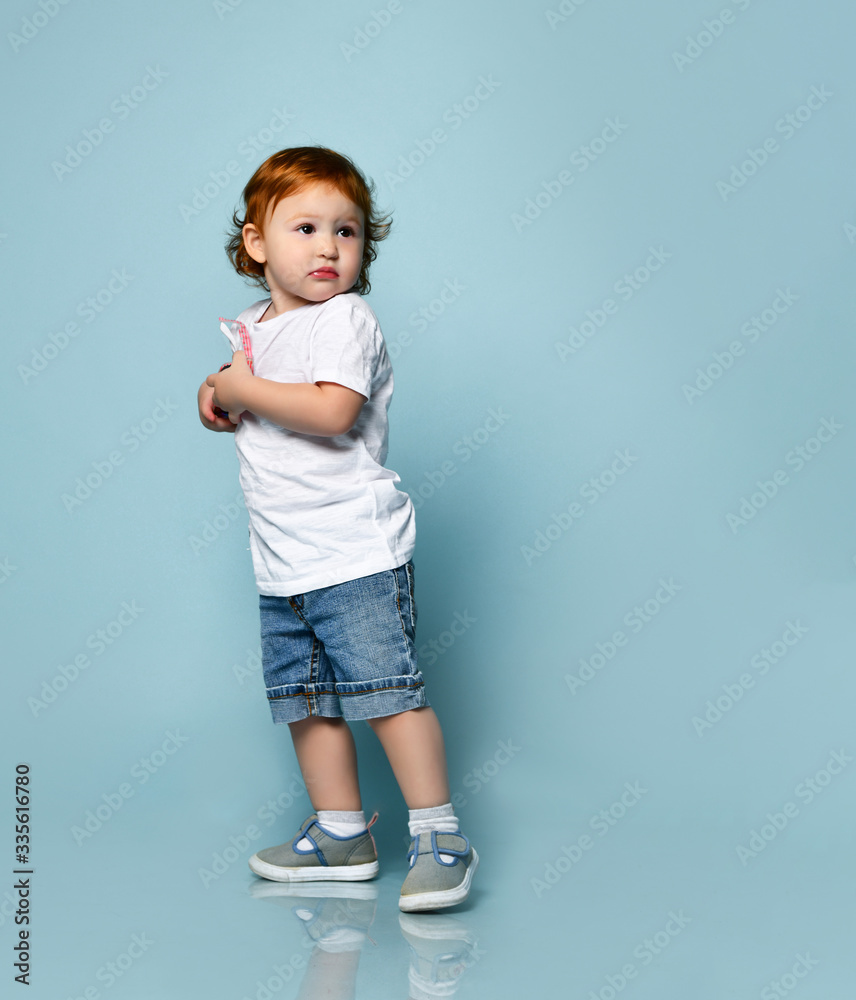 Ginger toddler boy or girl in white t-shirt, socks and shoes, denim shorts. Holding sunglasses, posing sideways on blue background
