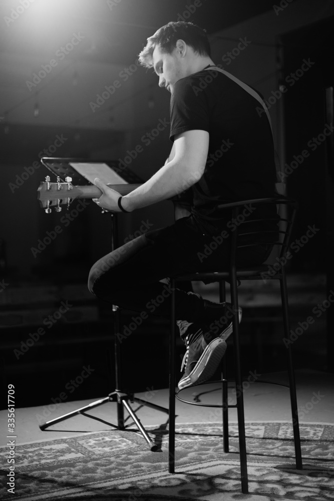 A young handsome guy of European appearance plays an acoustic guitar and sits on a chair. Black and white photography. A young handsome guy of European appearance plays an acoustic guitar and sits on a chair. Black and white photography.