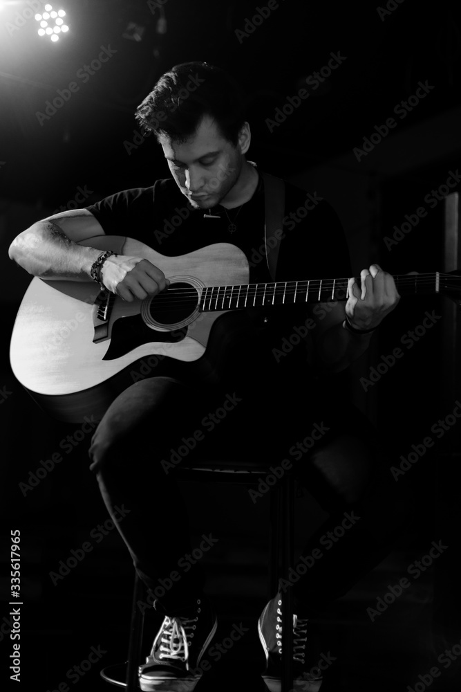 A young handsome guy of European appearance plays an acoustic guitar and sits on a chair. Black and white photography.