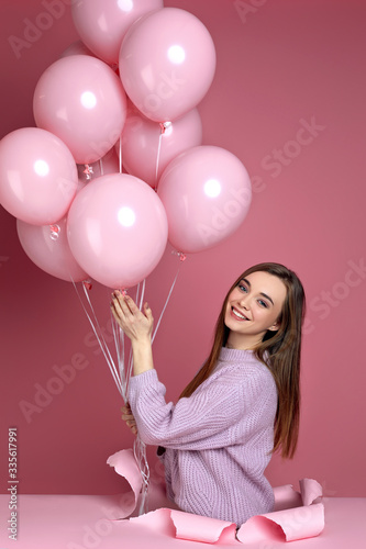 Smiling cute woman with pastel pink air balloons standing in torn paper hole on pink background. Beautiful happy young girl as a gift for birthday holiday.