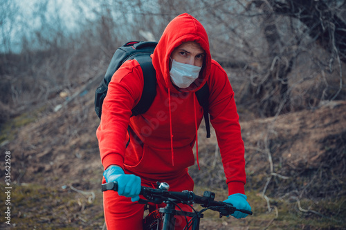 Young man in medical mask and gloves sitting on bicycle in countryside. Male protecting yourself from diseases on walk. Concept of threat of coronavirus epidemic infection.