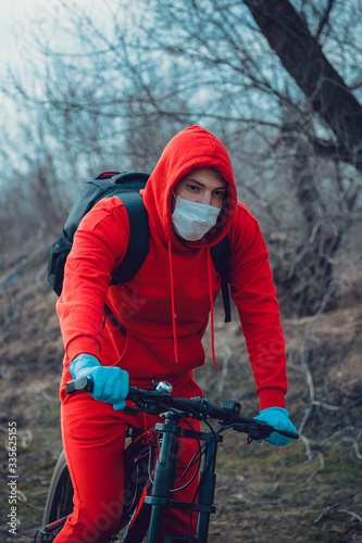 Young man in medical mask and gloves sitting on bicycle in countryside. Male protecting yourself from diseases on walk. Concept of threat of coronavirus epidemic infection.