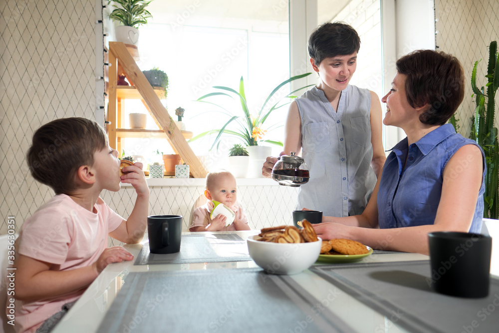 A young homosexual lesbian family with two children, a boy and a girl, at home in the kitchen forgiving the windows drinking tea and socialize.