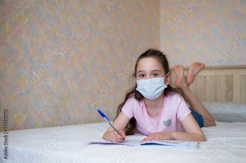 little girl in a protective mask writes in a notebook during quarantine. Distance learning education. look at camera