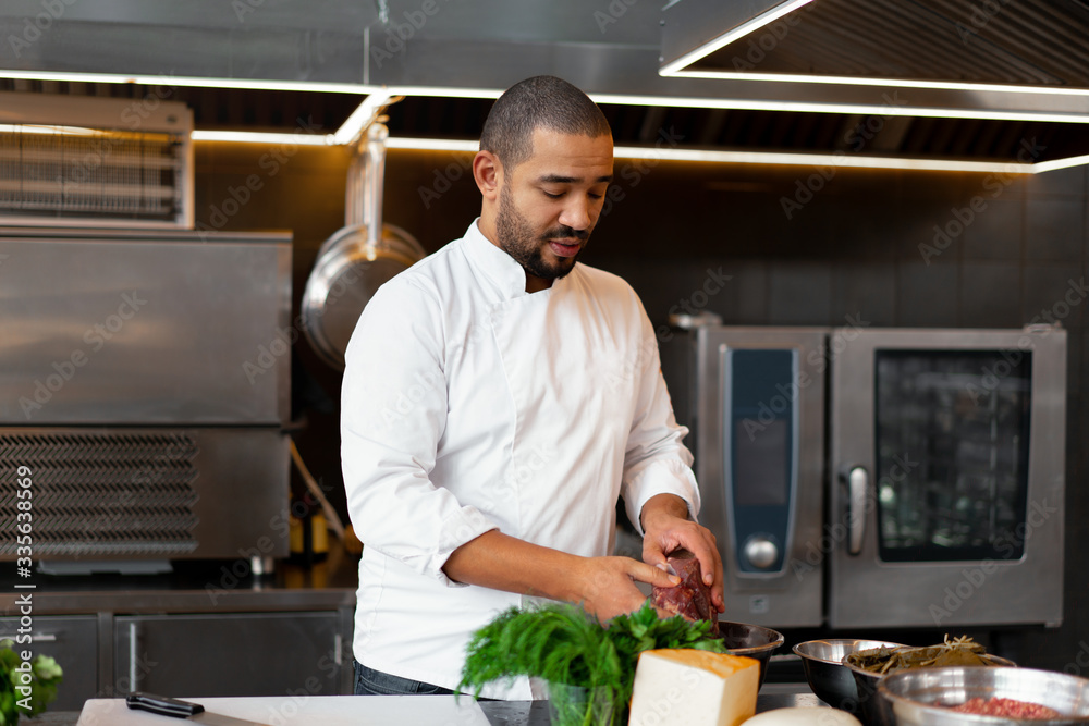 Handsome young African chef standing in professional kitchen in restaurant preparing a meal of meat and cheese vegetables. Handsome young African chef standing in professional kitchen in restaurant preparing a meal of meat and cheese vegetables.