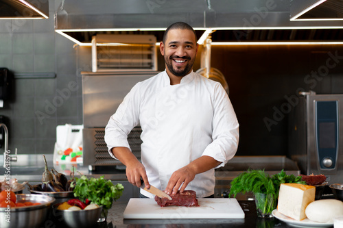 Handsome young African chef standing in professional kitchen in restaurant preparing a meal of meat and cheese vegetables.