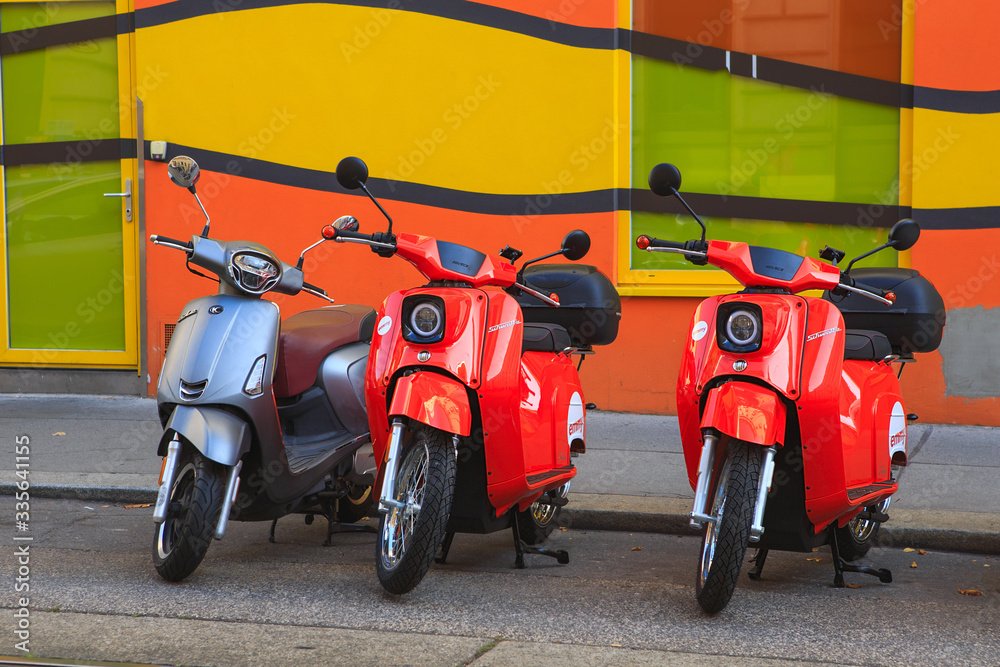 An electric scooters of the brand "Schwalbe" and Kymco Like stand at the  roadside. Emmy electric scooters for sharing. Red bikes against bright  background Photos | Adobe Stock