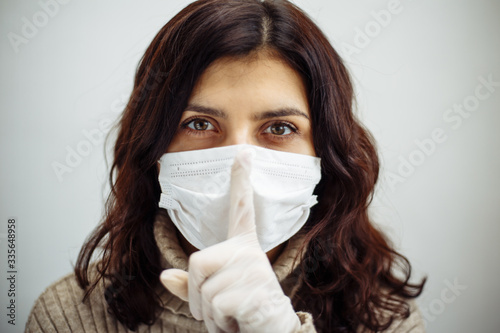 Portrait of young woman holds on her finger showing silence sign and has to stay home during quarantine due to coronavirus pandemia. Beautiful girl wearing medical mask and gloves. Covid-19 epidemia.